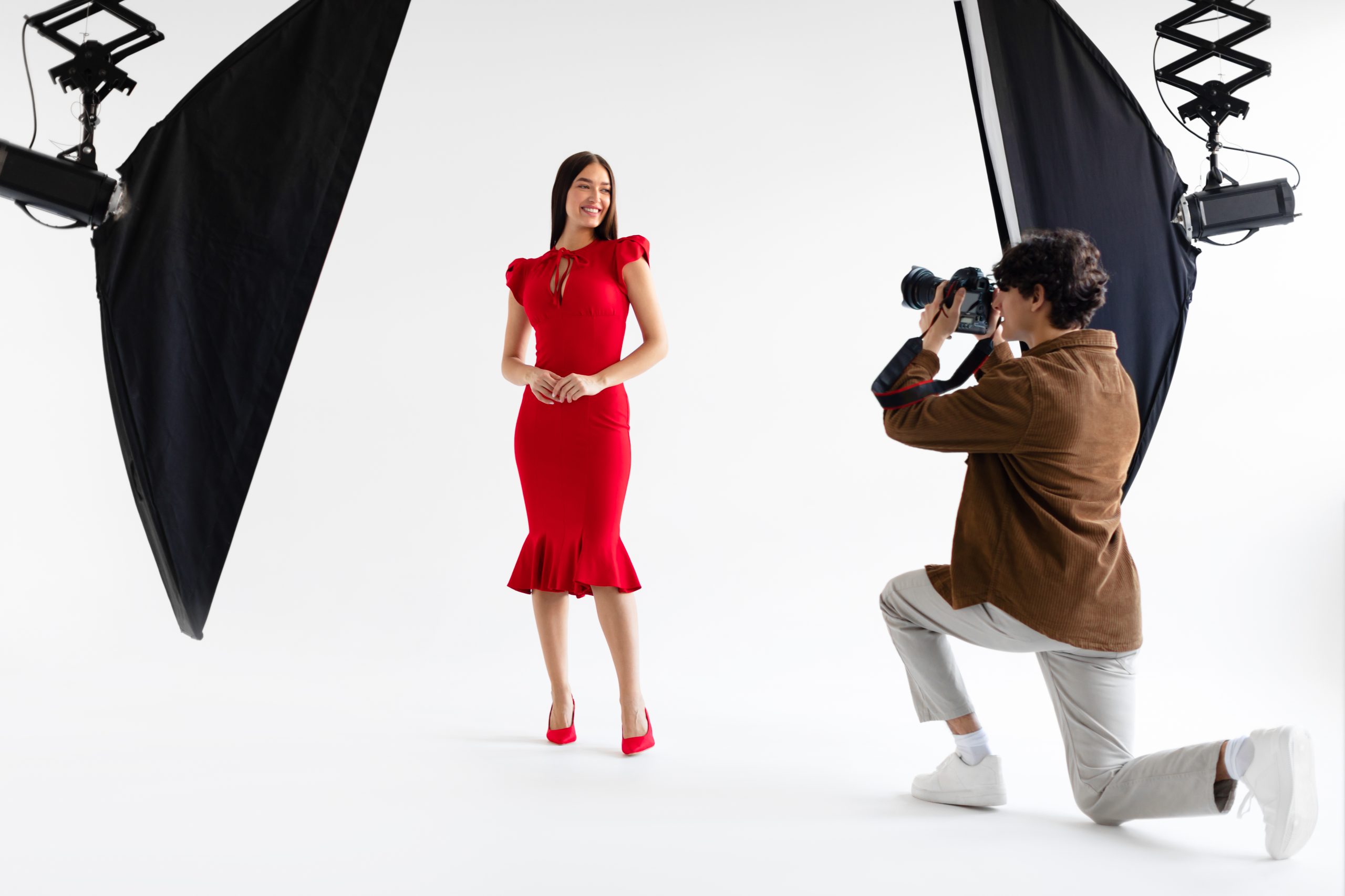 Séance photo de mode. Photographe professionnel prenant des photos d'un beau modèle féminin en robe rouge élégante, posant sur fond blanc dans un studio photo avec un équipement d'éclairage moderne
